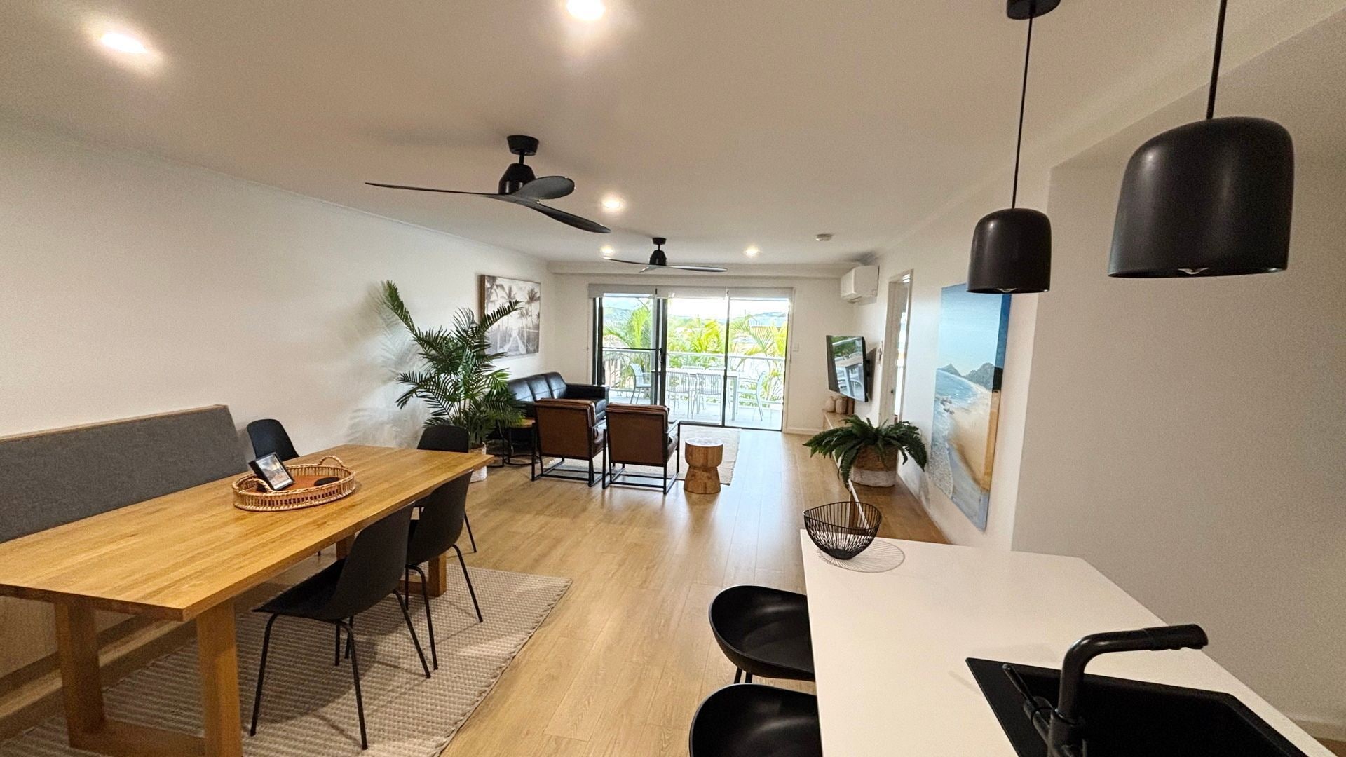 A modern dining area with a wooden table, black chairs, a ceiling fan, and a view of the outdoors through a glass door.
