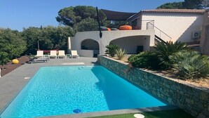 Une piscine rectangulaire avec un mur en pierre et une zone herbeuse à l’avant. On aperçoit des chaises longues et un espace de repos couvert à l’arrière-plan.