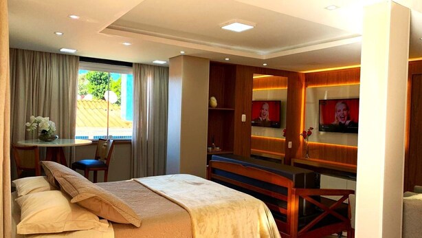 A hotel room with a bed, a desk, a TV, and a window with curtains. - Loft Vale das Montanhas - Gramado - RS (Gramado)