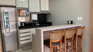 A modern kitchen with a stainless steel refrigerator, white cabinets, a black countertop, and a small island with three wooden bar stools. - Loft Vale das Montanhas - Gramado - RS (Gramado)