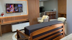 A modern living room with a flat-screen TV, wooden furniture, and a kitchen in the background. - Loft Vale das Montanhas - Gramado - RS (Gramado)