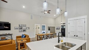 A modern kitchen with a large island, a dining area with a wooden table and chairs, a living room with a sofa and a TV, and a hallway leading to bedrooms. - The Turn at Kilmarlic - Kilmarlic Golf Club, Sits on 10th Tee, Water Park Nearby, YR Flex Stays (Powells Point)