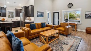 A spacious living room with a large brown leather sofa, a wooden coffee table, and a patterned rug. There is a kitchen area with dark cabinets and a refrigerator. A dining area with a wooden table and chairs is visible. - The Turn at Kilmarlic - Kilmarlic Golf Club, Sits on 10th Tee, Water Park Nearby, YR Flex Stays (Powells Point)