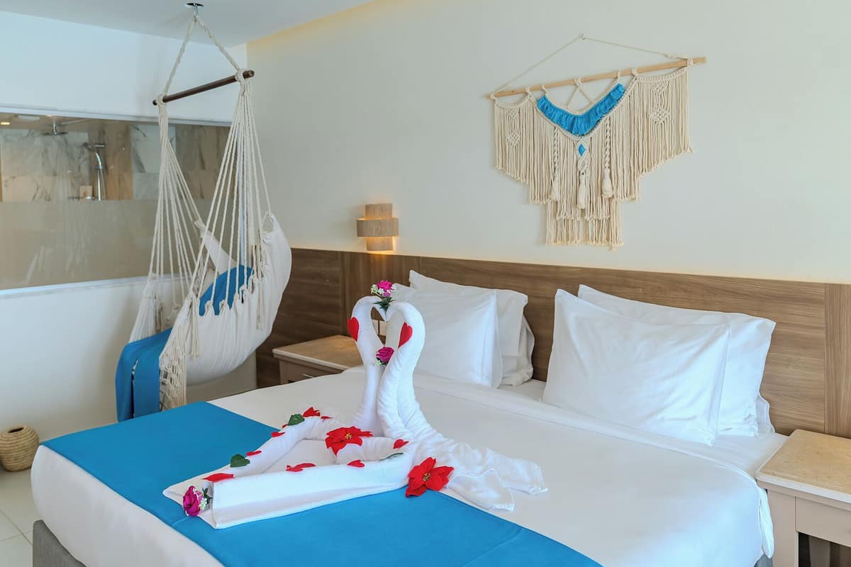 Quarto de hotel com cama, cadeira suspensa e um painel de macramé na parede.