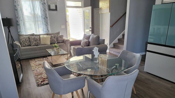 Een moderne woonkamer met een glazen eettafel, grijze banken en een trap naar een ander niveau.