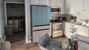 Een moderne keuken met witte kasten, een blauwe koelkast, roestvrijstalen apparatuur en een glazen eettafel met een stoel.