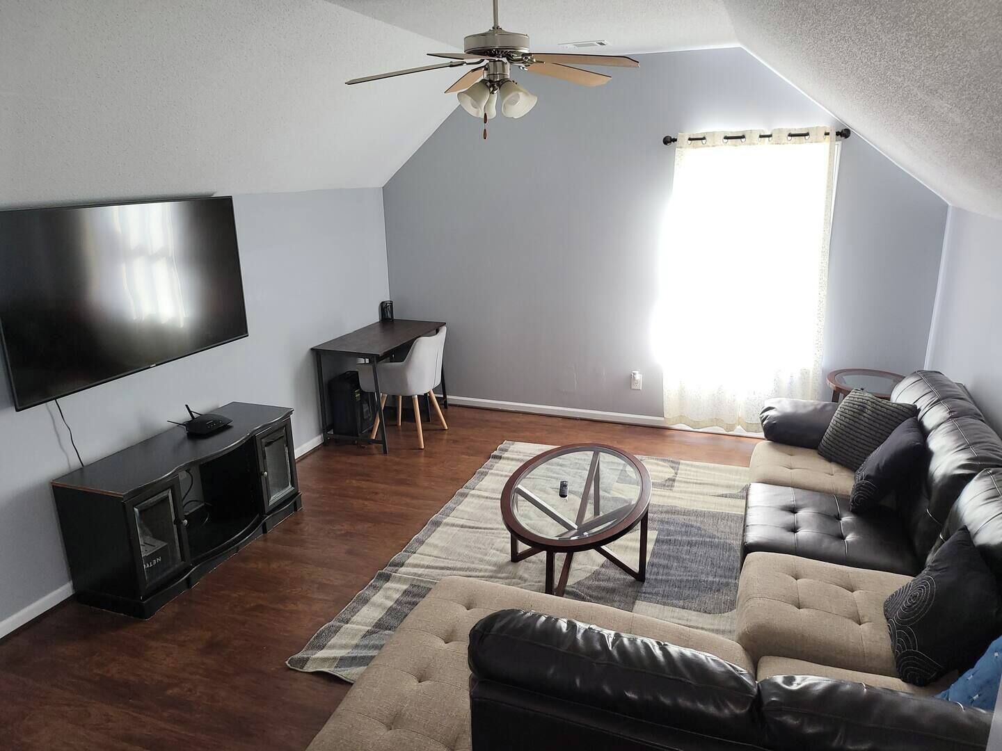 A living room with a flat-screen TV, a dark wooden entertainment unit, a leather sofa, a glass coffee table, and a ceiling fan.