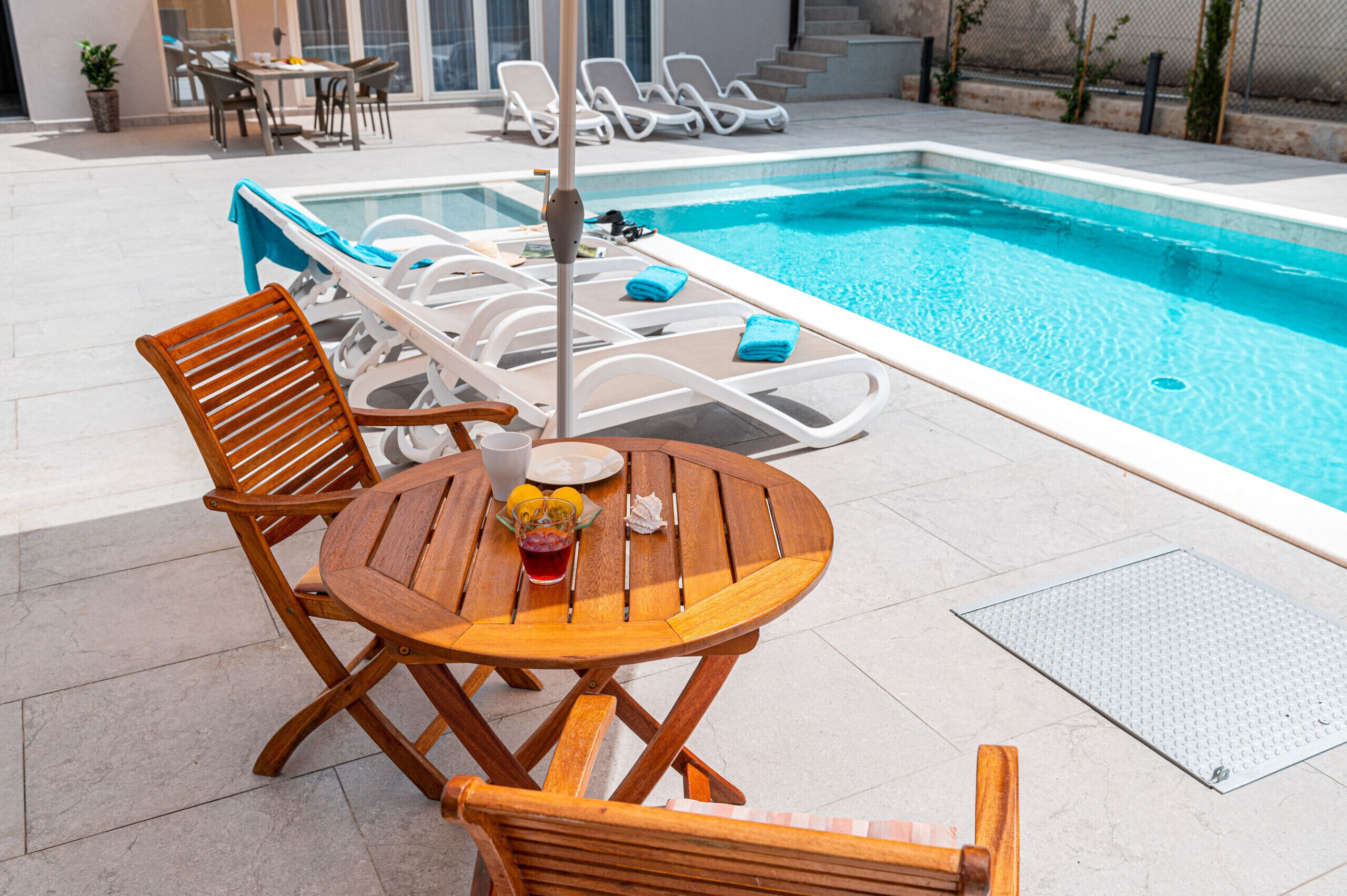 A poolside area with a wooden table and chairs, a pool, and lounge chairs.
