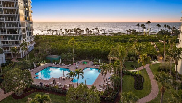 A pool area with a view of the ocean, surrounded by palm trees and greenery. - Modern Condo Steps from the Private Beach with Amazing Views (Naples)