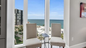 A room with two chairs facing a large window overlooking a beach and cityscape. - Modern Condo Steps from the Private Beach with Amazing Views (Naples)
