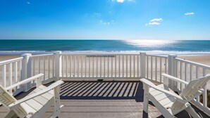 Blick auf einen Strand mit einer Holzterrasse, weißen Geländern und einem Schild, das darauf hinweist, dass Sitzen oder Lehen an der Brüstung verboten ist.
