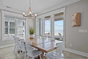 A dining area with a wooden table and white chairs, large windows with blinds, and a view of the ocean.