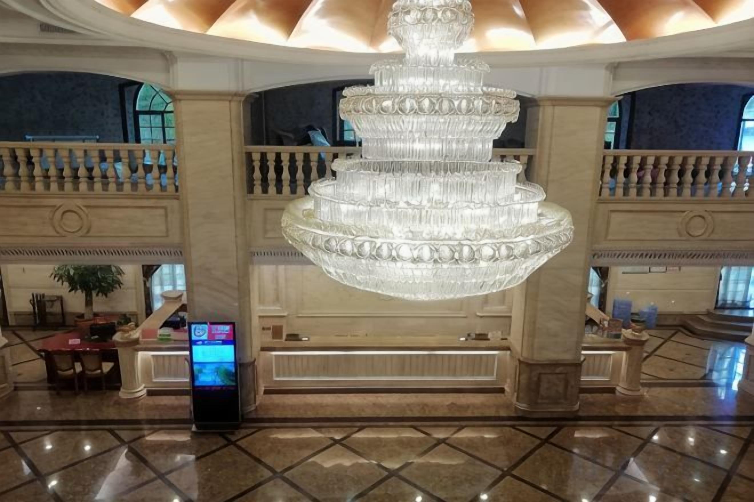 A lobby with a large, multi-tiered crystal chandelier, marble flooring, and a reception desk with a digital display.