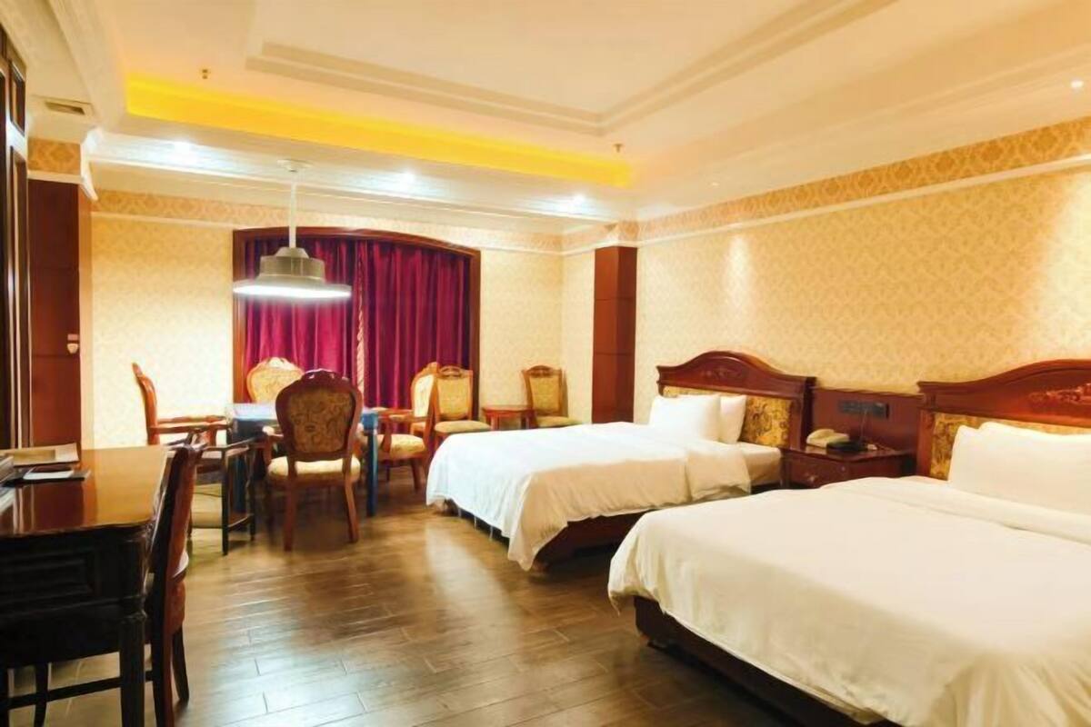 Une chambre d’hôtel avec deux lits, une table à manger, des chaises, un lustre et un rideau rouge.