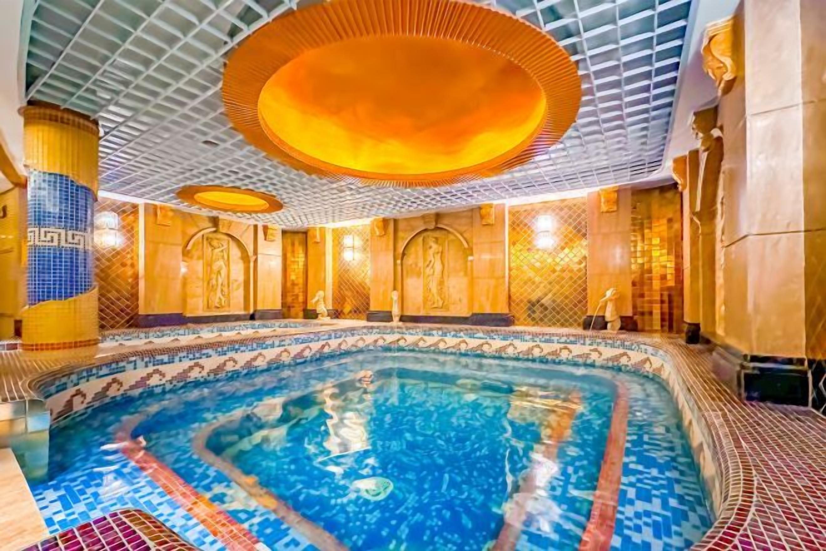 An indoor swimming pool with a tiled floor, a central pool area, and a ceiling with a geometric pattern.
