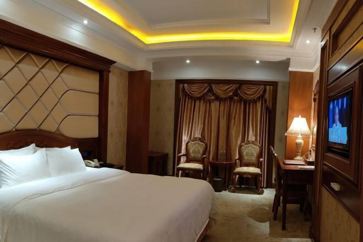 Une chambre d’hôtel avec un grand lit, un bureau avec une lampe, deux chaises, une télévision et un rideau.