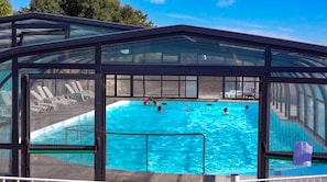 A covered swimming pool with clear blue water, surrounded by lounge chairs and a glass enclosure. - Camping le Dolmen (Carnac)