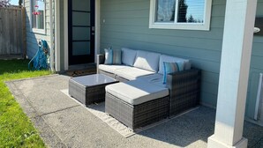 Eine überdachte Veranda mit einem grauen Rattan-Sofa und zwei Rattan-Tischen, einem Holzzaun und einer blauen Tür.