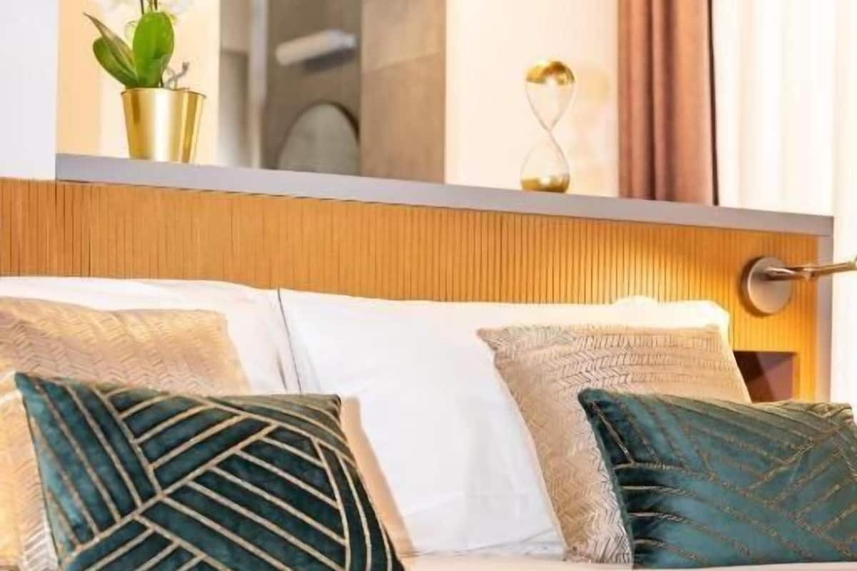 Ein Hotelzimmer mit einem Bett, zwei Zierkissen, einer grünen Tagesdecke, einer Vase mit weißen Blumen und einem Spiegel über dem Bett.