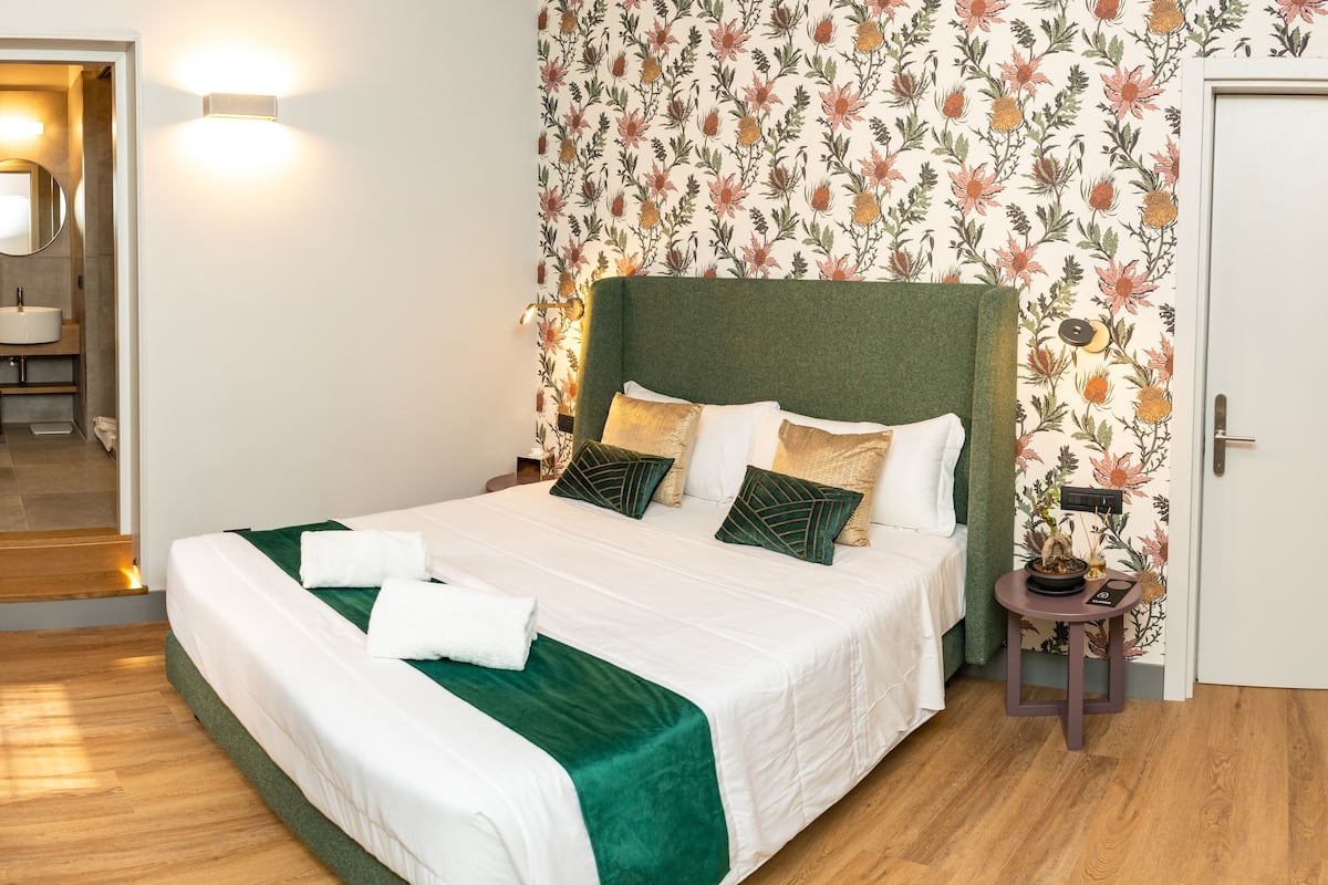 Ein ordentlich bezogenes Bett mit einem grünen Kopfteil, weißer Bettwäsche und dekorativen Kissen. Ein kleiner Nachttisch mit Lampe und Dekoration ist zu sehen. Der Raum hat floralen Wandbelag und Holzdielen.