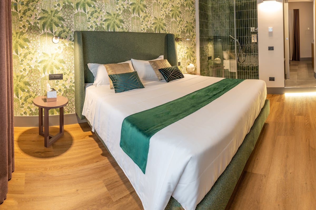 Ein Hotelzimmer mit einem großen Bett, einem kleinen Nachttisch, einem grünen Kopfteil und einem durch eine Glastür sichtbaren Duschbereich.