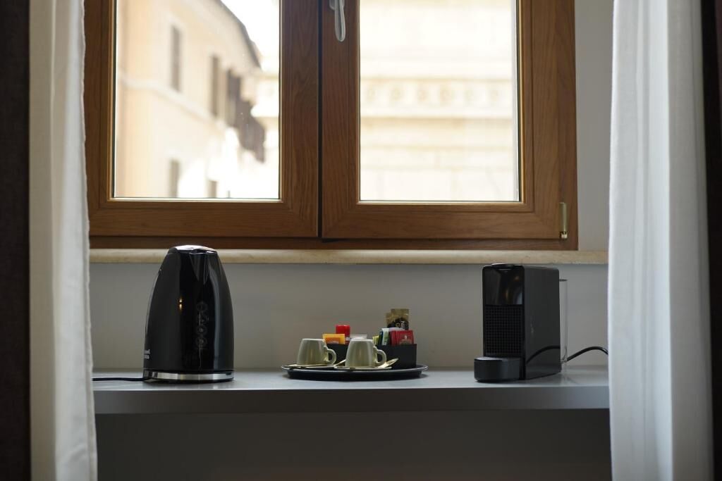Una ventana con marco de madera, una cafetera de café negro, dos tazas blancas y un altavoz negro sobre un estante blanco.