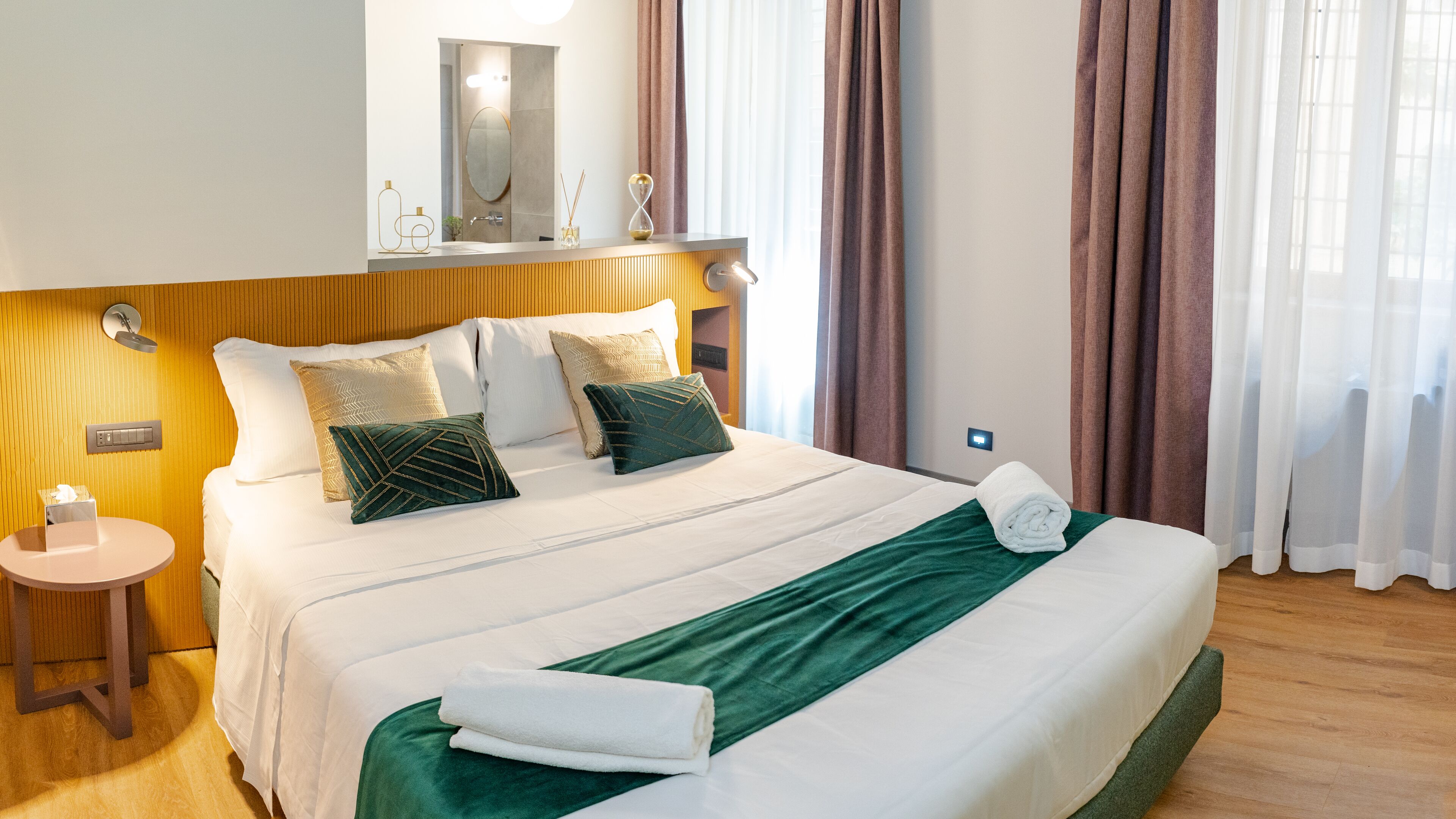 Una cama bien tendida con un plaid verde, una mesita de noche con una caja de pañuelos y una lámpara de pared.