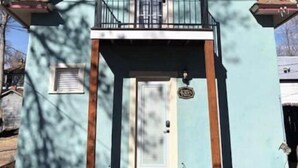 A small, single-story building with a light blue exterior, a front door, a small balcony with a metal railing, and a visible mailbox. - Cozy Carriage House - Downtown (Colorado Springs)