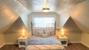 A bedroom with a sloped ceiling, a bed with a headboard, two bedside tables, and a window with blinds. - Cozy Carriage House - Downtown (Colorado Springs)