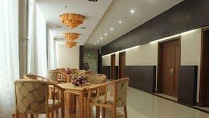 A modern hotel lobby with a wooden dining table, chairs, and a floral centerpiece. There are wooden doors and a wall-mounted light fixture.
