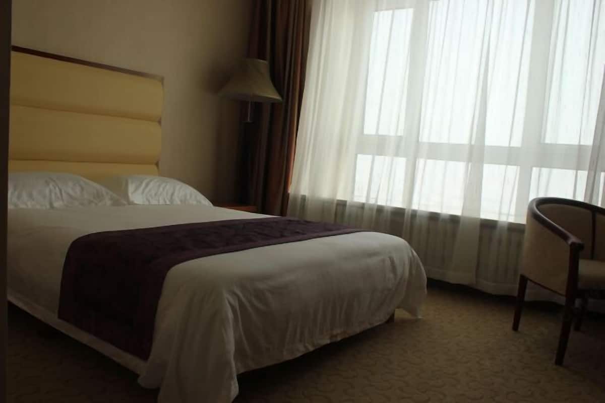 Een hotelkamer met een groot bed, een stoel en een raam met dunne gordijnen.