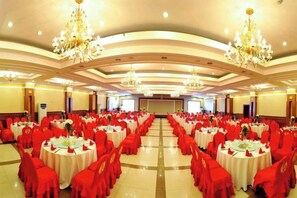 Une salle de banquet avec des tables rondes et des chaises rouges, des lustres et une scène à l’avant.