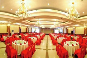 A banquet hall with round tables and red chairs, chandeliers, and a stage at the front.