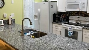 Una cucina con piani di lavoro in granito, mobili bianchi, lavello in acciaio inossidabile, frigorifero, microonde, fornello e una sveglia appesa al muro.