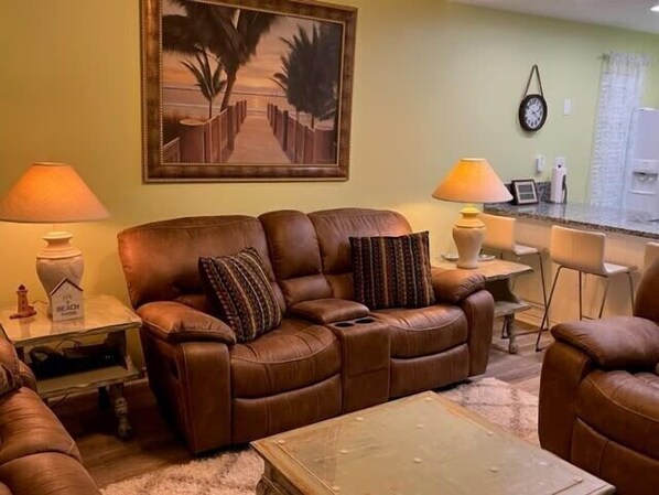 A living room with a brown leather sofa, two side tables with lamps, a coffee table, and a painting of a beach scene.