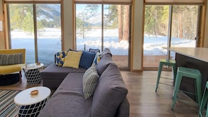 Un accogliente soggiorno con un divano grigio, cuscini decorati, una poltrona gialla e un tavolino rotondo. Ampie finestre offrono vista su un paesaggio innevato e alberi.