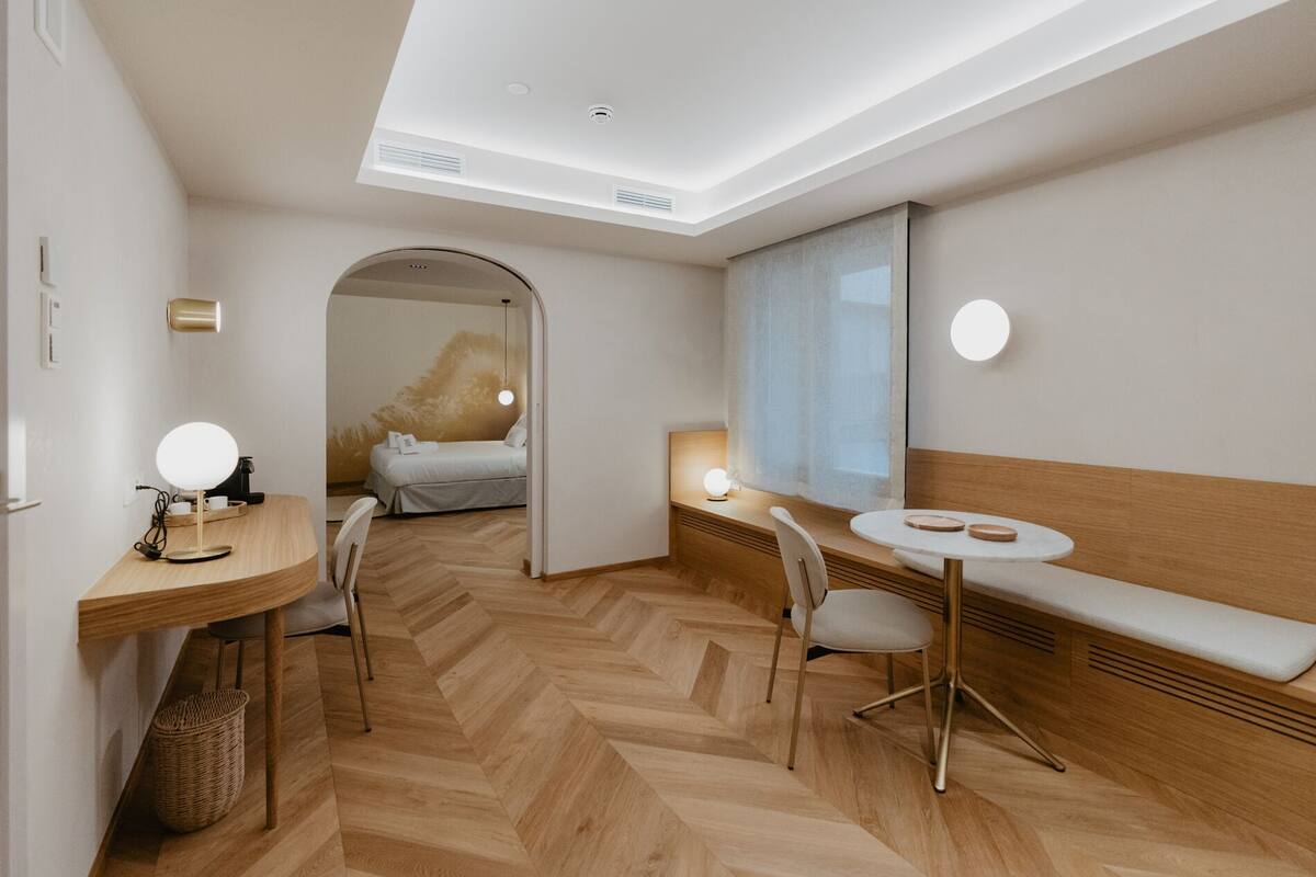Een moderne hotelkamer met een visgraat parketvloer, een houten bureau met lamp, een nachtkastje met lamp, een bankje en een groot raam met uitzicht.