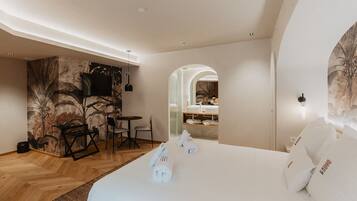 Una habitación de hotel moderna con una cama grande, una zona de comedor con sillas, una mesita y un baño visible a través de un arco.