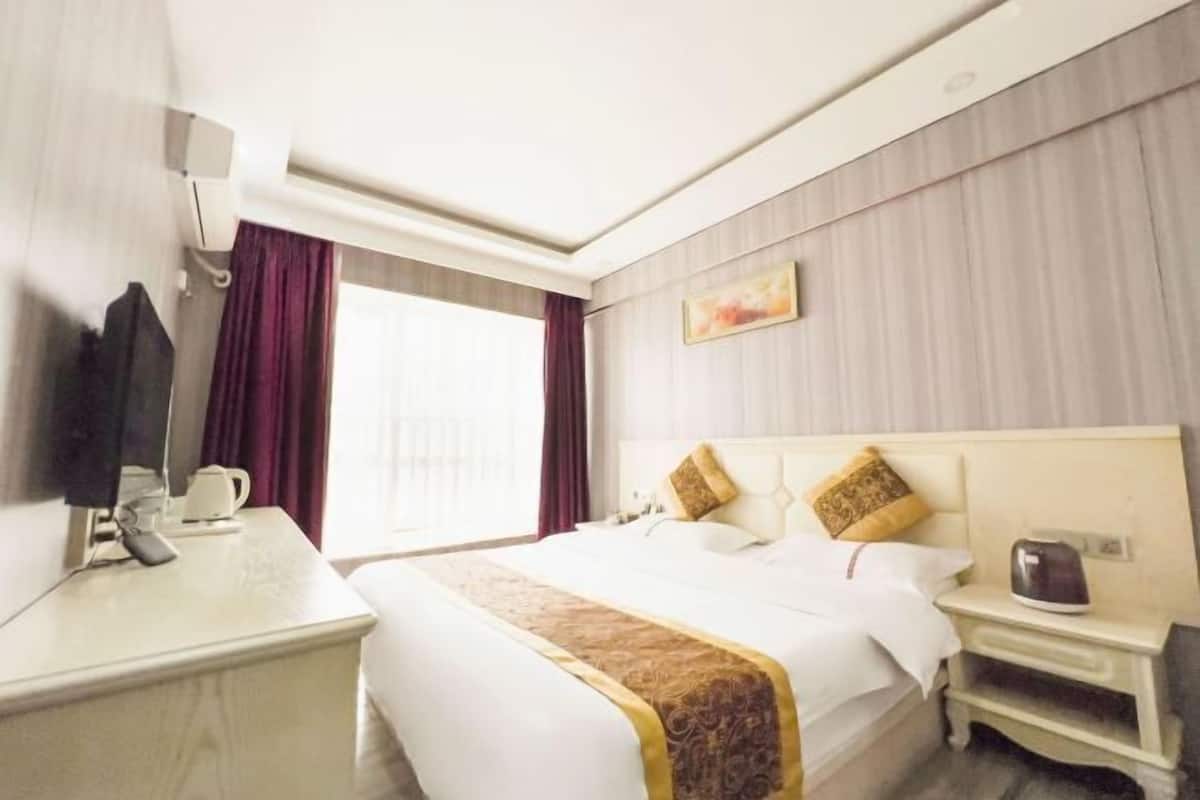 Ein Hotelzimmer mit einem großen Bett, einem Nachttisch, einem Schreibtisch mit Stuhl, einem Fenster mit Vorhängen und einem Bild an der Wand.