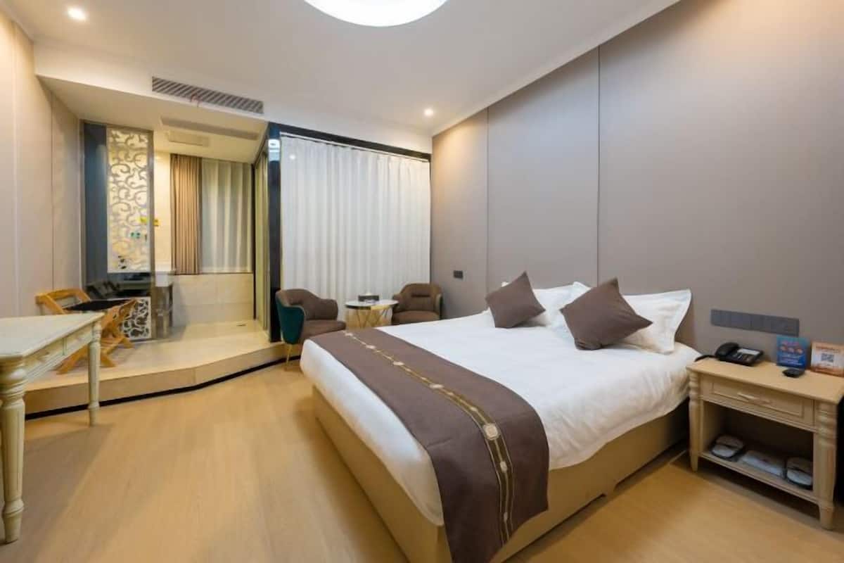A hotel room with a large bed, a desk, a chair, a small table, and a bedside table with a lamp.
