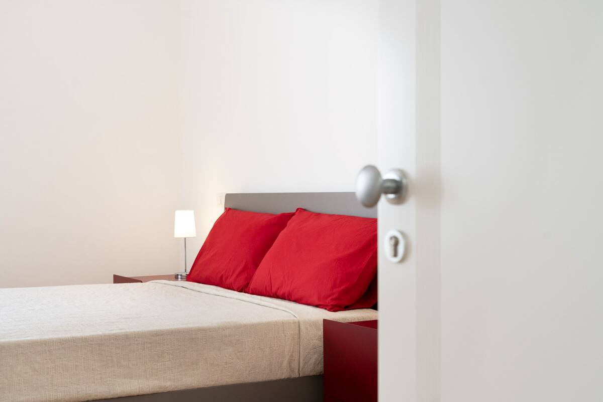 A bed with a beige bedspread and two red pillows, a bedside table with a lamp, and a door with a keyhole.