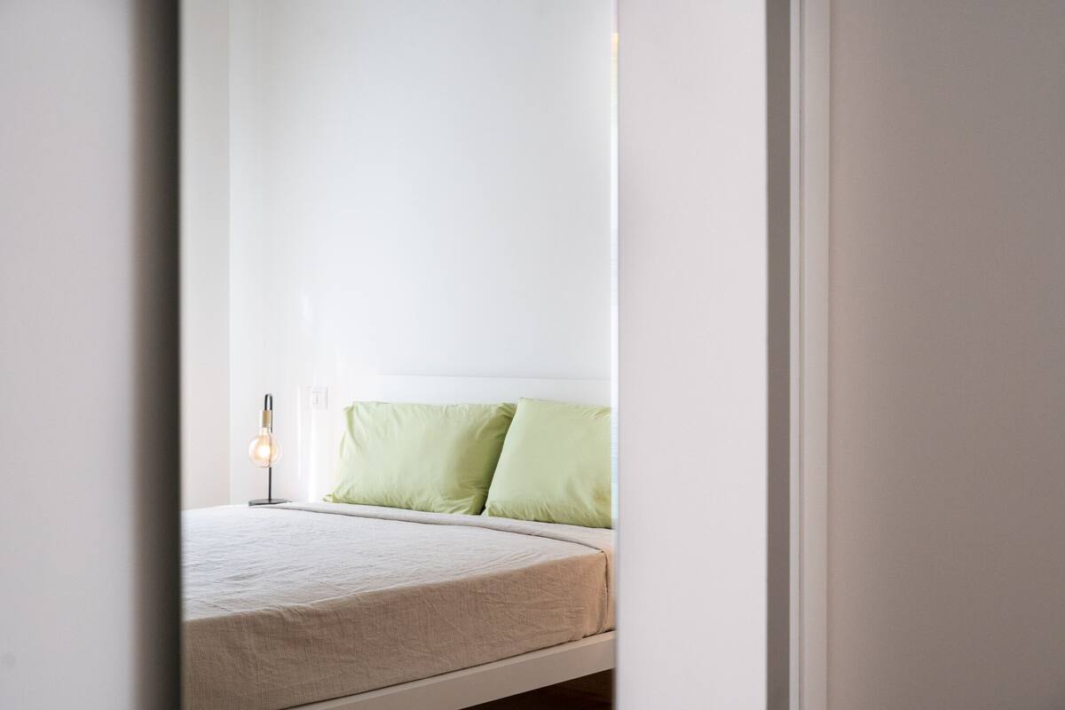 A bed with a white mattress and green pillows, a bedside lamp, and a white wall.