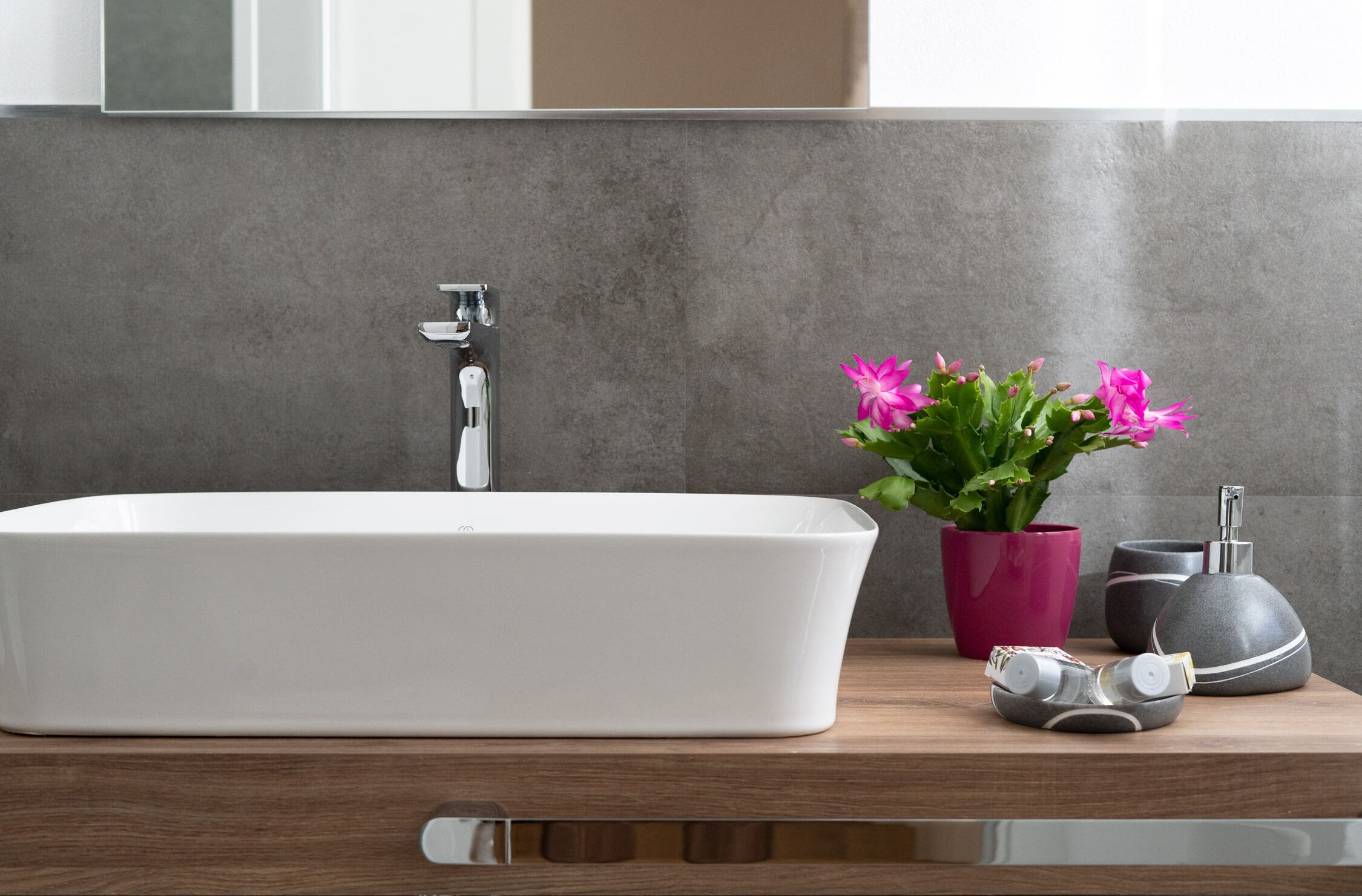 Une salle de bain avec un lavabo blanc, un vase rose contenant des fleurs et un distributeur de savon gris.