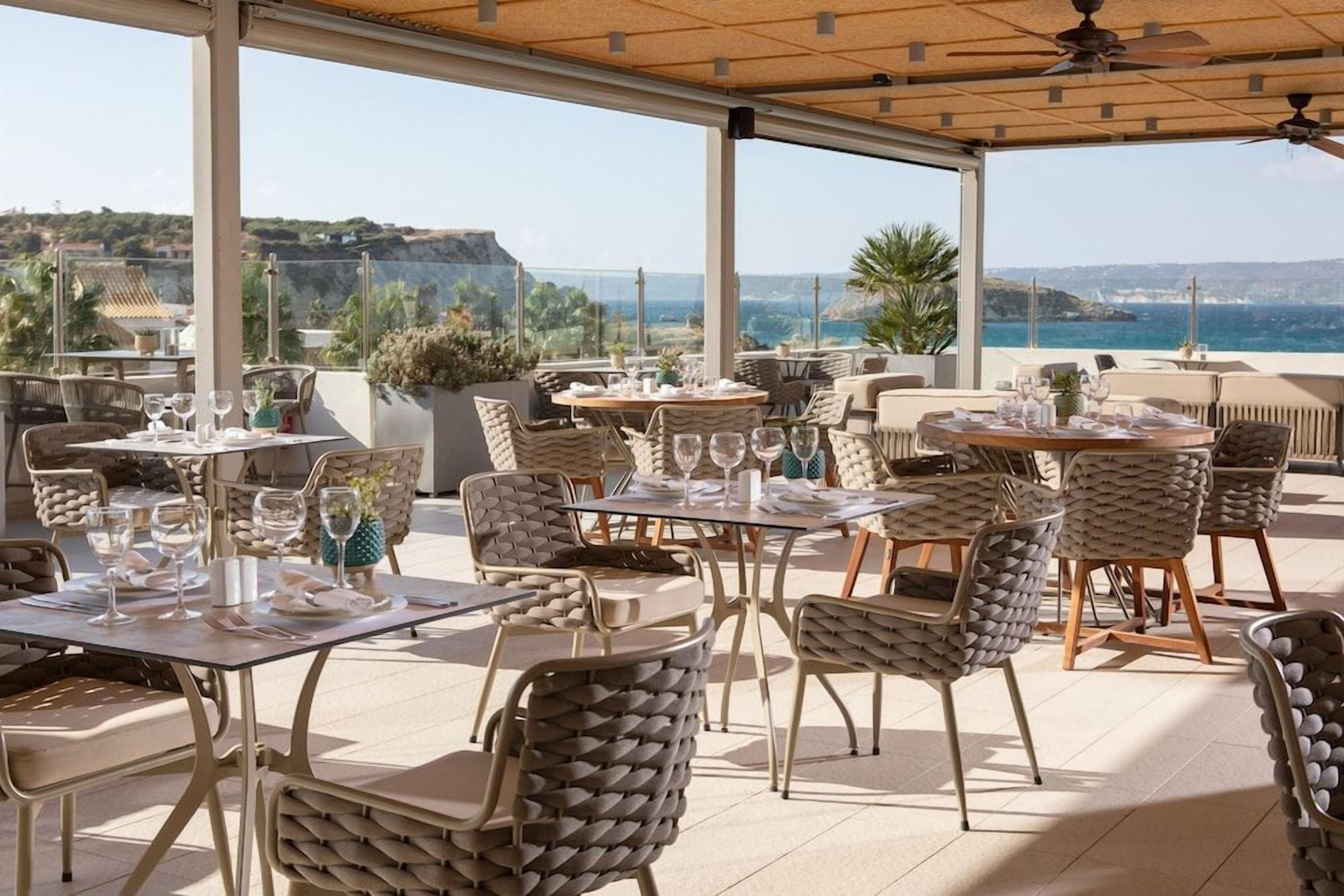 An outdoor dining area with tables and chairs, a view of the sea, and a building in the background.