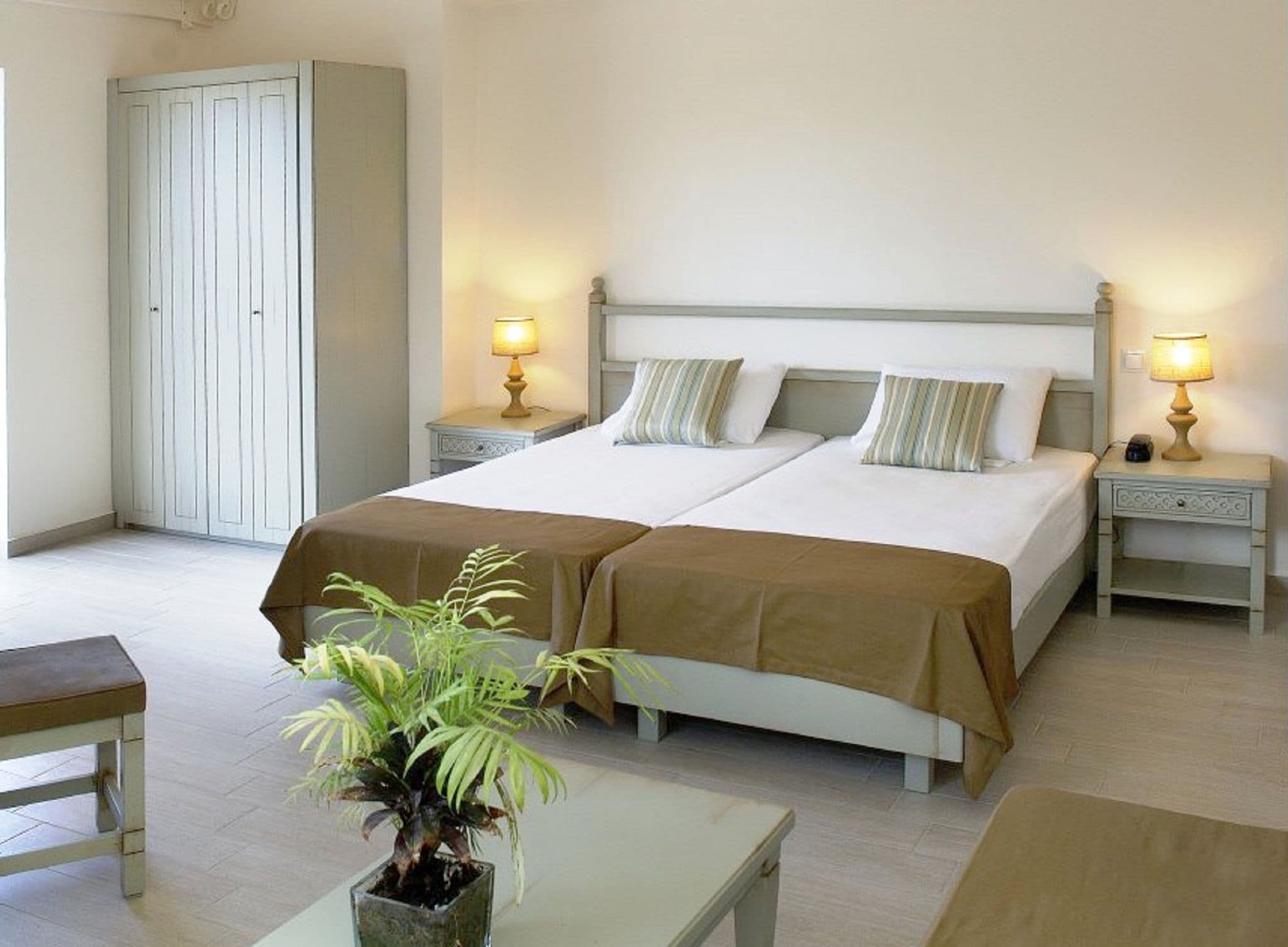 Uma cama de casal com duas mesas de cabeceira, um criado-mudo, uma planta em vaso e um guarda-roupa.