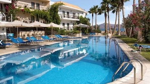 A swimming pool with lounge chairs and umbrellas, surrounded by palm trees and a building with balconies. - Almyrida Beach Hotel (Apokoronas)