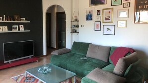 A living room with a green sofa, a glass coffee table, and a flat-screen TV. There are framed pictures on the wall and a bookshelf with decorative items. - Penthaus am Luganersee - Comer See (Carlazzo)