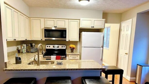 A kitchen with white cabinets, a stainless steel microwave, a black stove, a white refrigerator, and a granite countertop. There are three black barstools at the island.