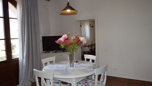 Ein Essbereich mit einem runden Tisch, weißen Stühlen, einer Blumenvase, einem Fernseher und einer Pendelleuchte.