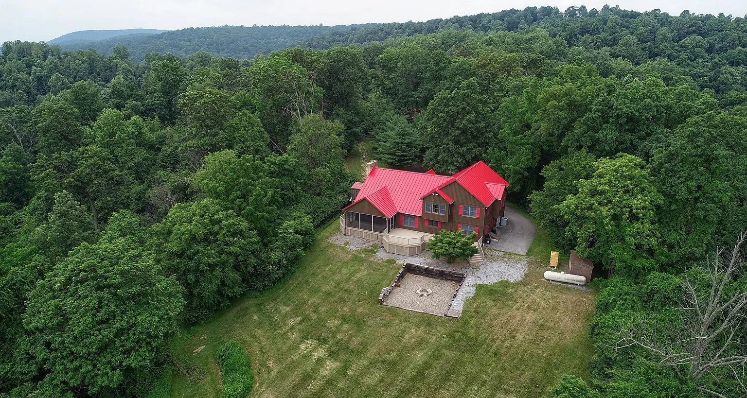 An aerial view of a house with a red roof surrounded by dense forest.